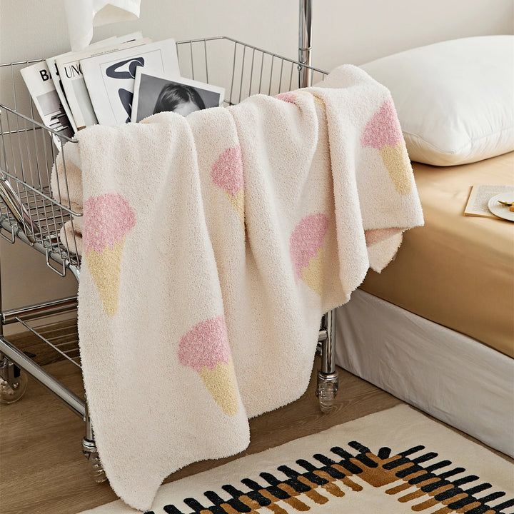 Ice cream pattern blanket hanging over a wire cart, showing its cream plush fabric and pink motifs