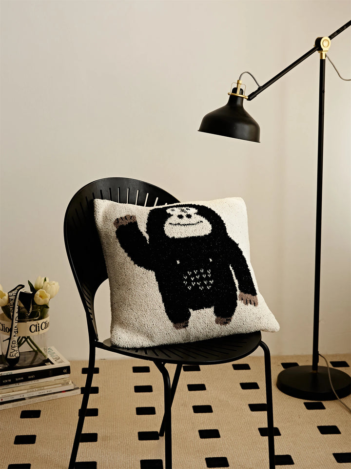 Cream Kawaii Gorilla pillow with a waving black gorilla displayed on a black chair beneath a floor lamp