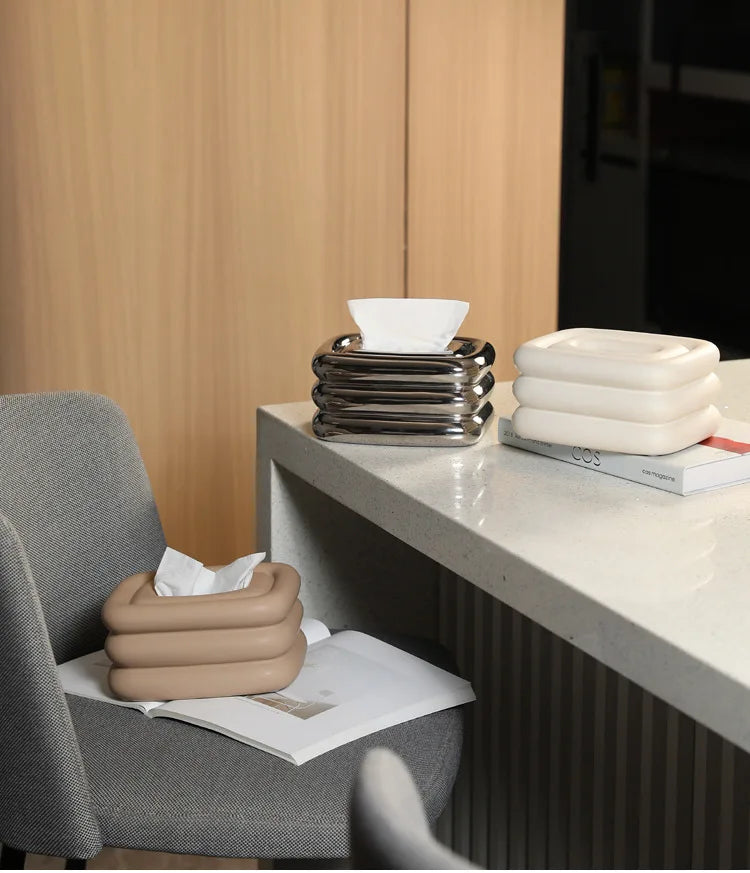 Three square tissue holders in silver, white, and light brown, each with rounded stacked sides.