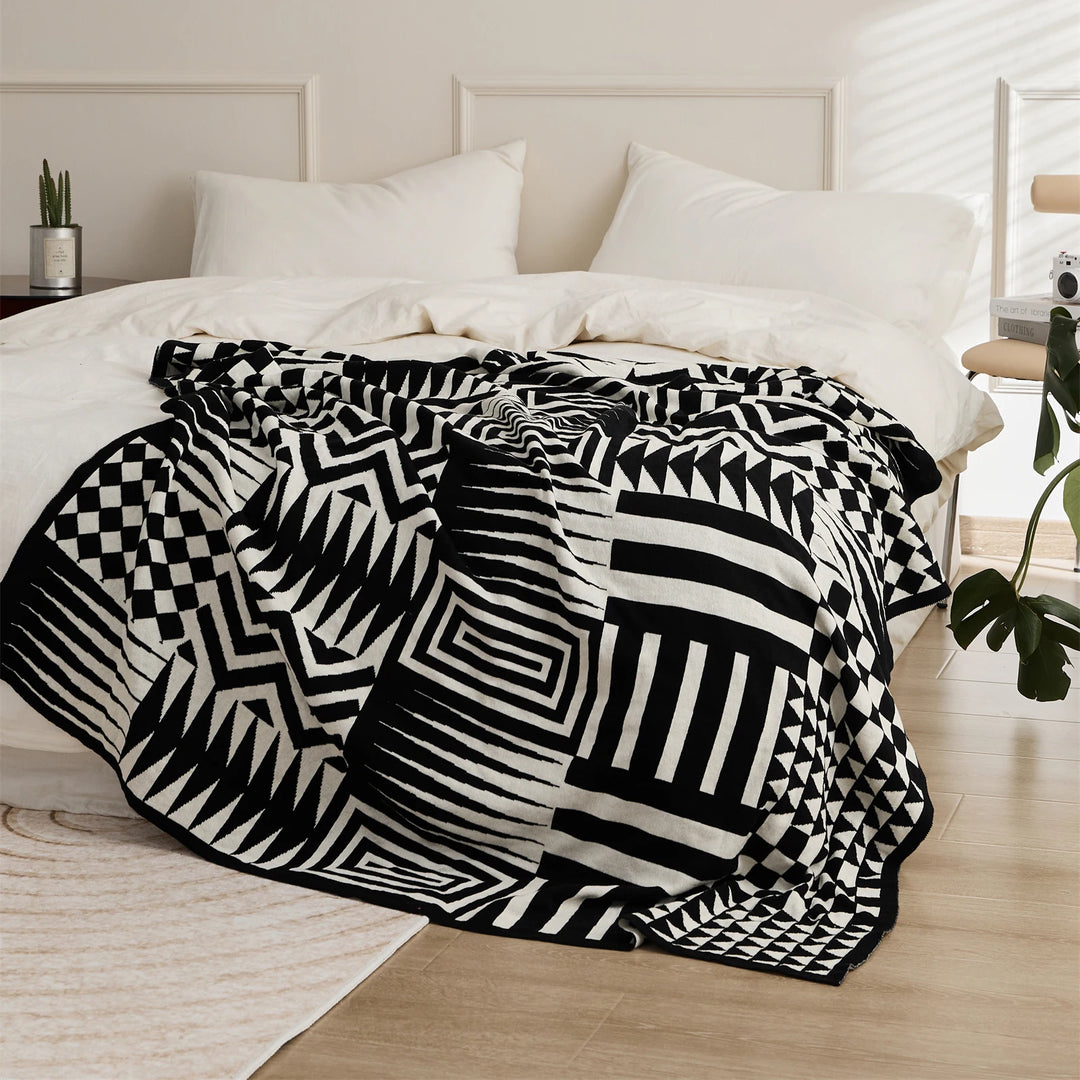 Black and cream geometric blanket spread across a bed, showing bold stripes, checks and angular motifs.